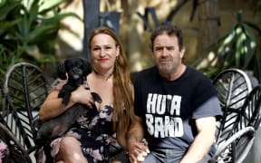 Elaine and Greg Exler sit on chairs holding hands and Elaine is holding their small dog.