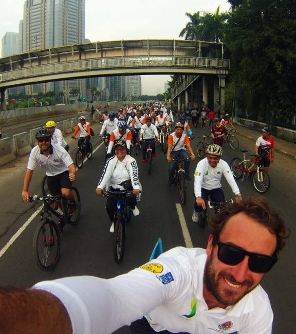 A man riding a bicycle wearing dark sunglasses and white lycra smiles as he takes a selfie, showing a huge cohort of people in white on bicycles riding down the road behind him.