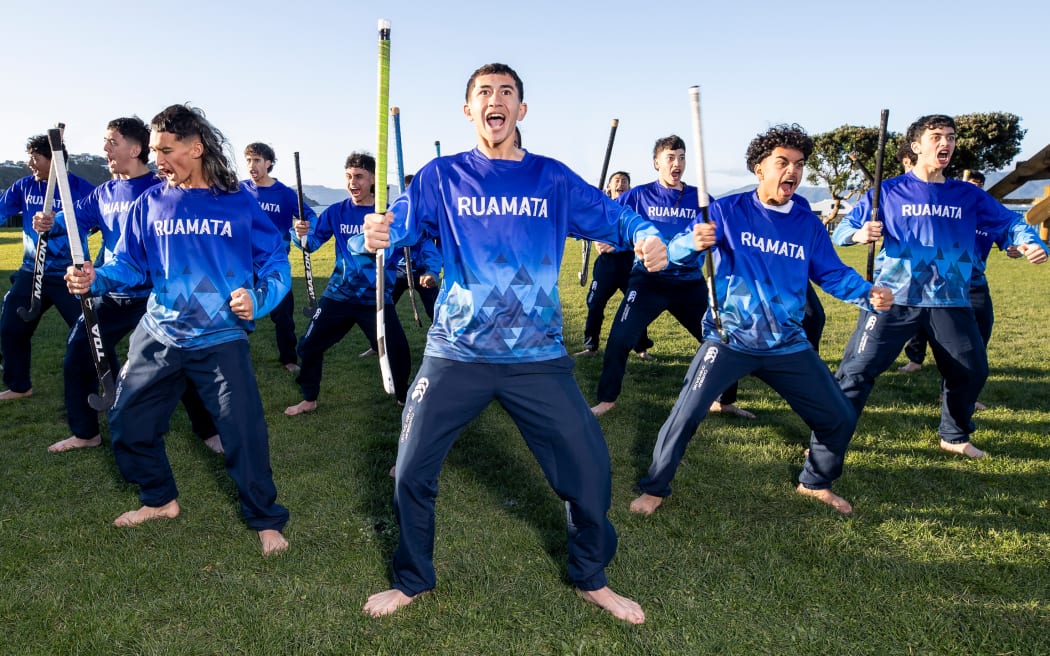 A group of teenagers in a blue sports uniform perform a haka holding hockey sticks.