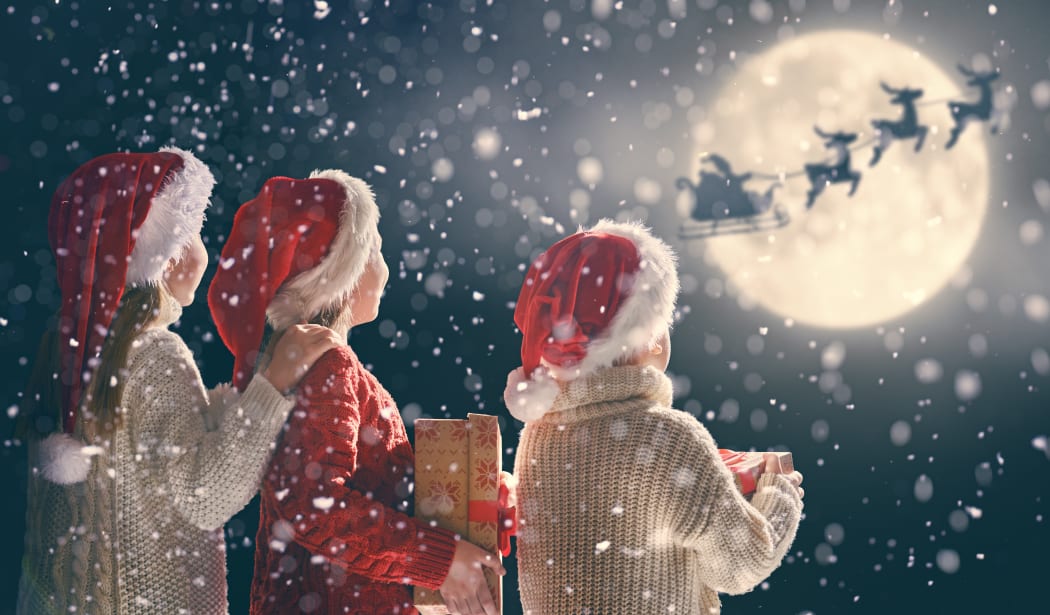 Merry Christmas and happy holidays! Cute little children with xmas presents. Santa Claus flying in his sleigh against moon sky. Kids enjoying the holiday with gifts on dark background.