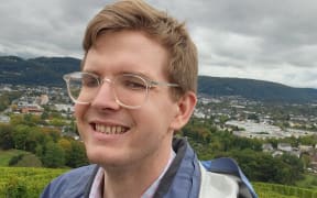 Henry Hickman is standing on the top of a hill and smiling into the camera. He is wearing a kind of greyish blue rain coat. The sky is very overcast, but it isn't raining.
