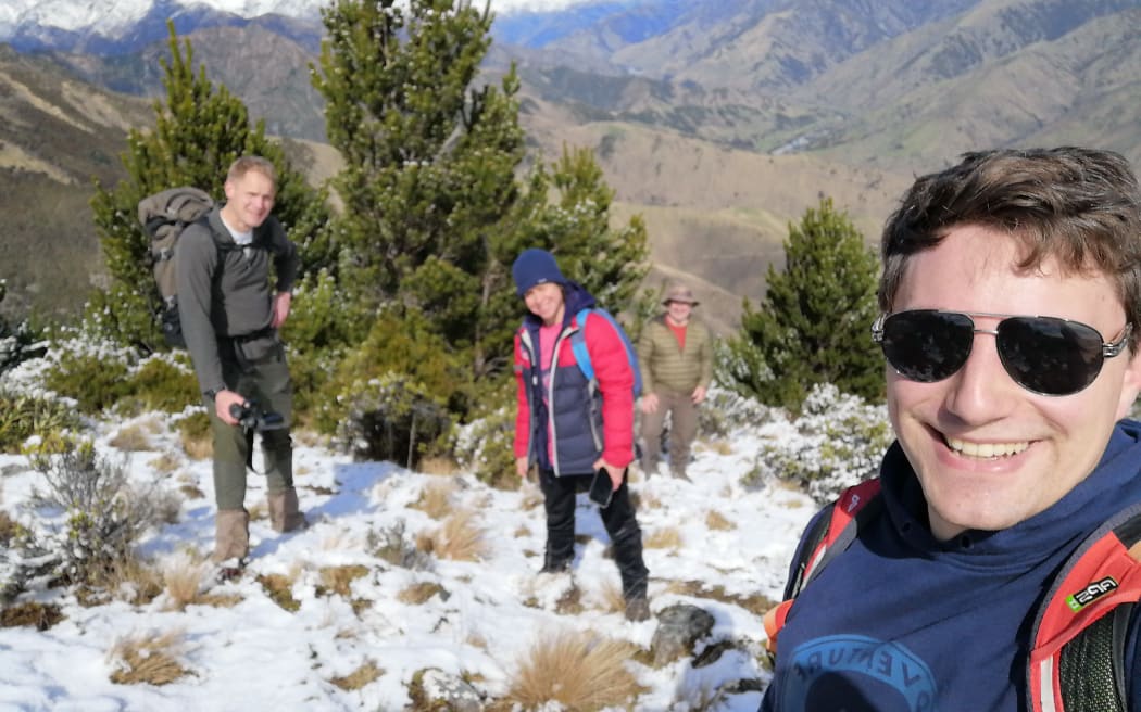 Dr Thomas Carlin is holding the camera and taking a selfie. He is in the bottom right-hand corner, wearing sunglasses. There are three people in the background standing in front of some wilding pines. There is snow on the ground, and they are clearly at the top of a hill. There is a mountain range behind them in the distance.