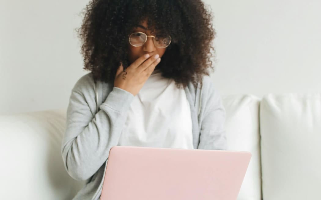 A woman wearing a white tshirt and grey cardigan covers her mouth with her hand in surprise as she looks at a laptop.ggen