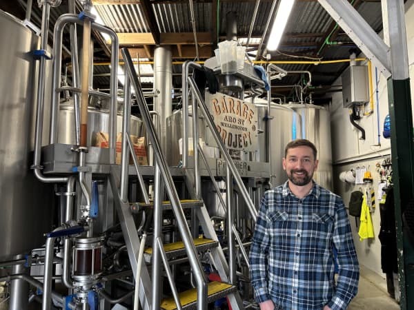A man in a blue flannel shirt stands in front of large brewery equipment in a warehouse, with a sign that says 'Garage Project' visible behind him.