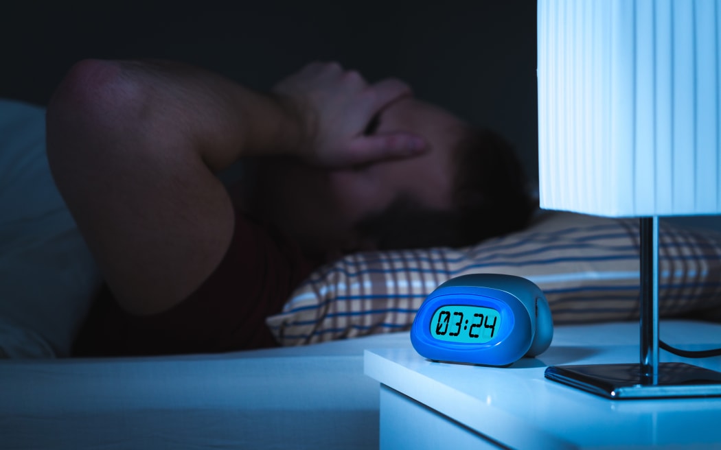 Insomnia and sleepless concept. Man unable to sleep. Exhausted and tired. Covering face with hand. Alarm clock on nightstand and bed in bedroom.