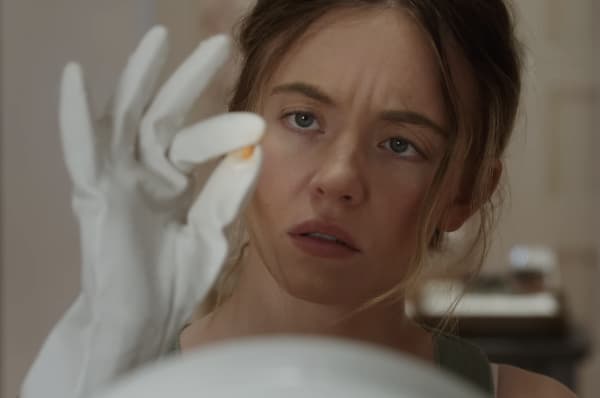 Sydney Sweeney, wearing white gloves, examines something small between two of her fingers.