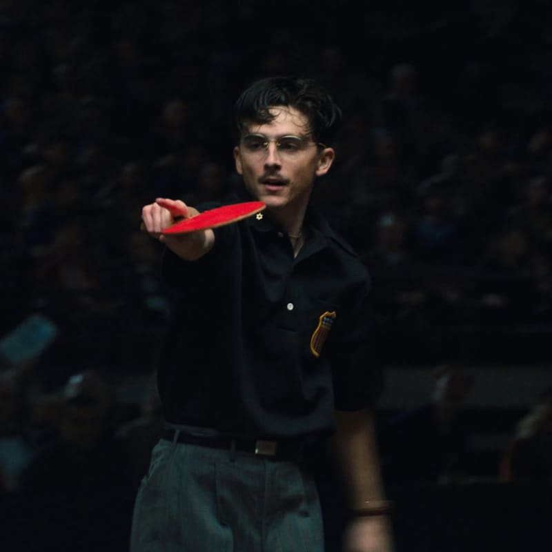 Timothee Chalamet, a young man with a moustache and glasses, holds a red ping-pong racquet as he points a finger.