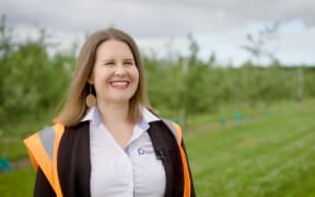 Emma Brown stands in an orchard. She has a big smile on her face. She is wearing a shirt, high vis, and big round bronze earings.
