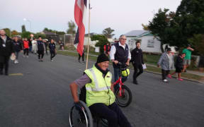 Wairoa resident Val Irwin is in a wheelchair and wears a yellow hi vis vest and black beanie. He has a tino rangatiratanga flag attached to the back of the wheelchair and other people walk alongside and behind him.
