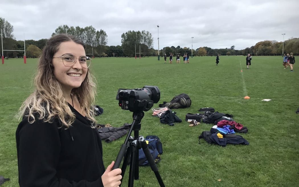 A woman with long blond hair smiles at the camera while standing behind a video camera on a tripod, pointed at a sports field.