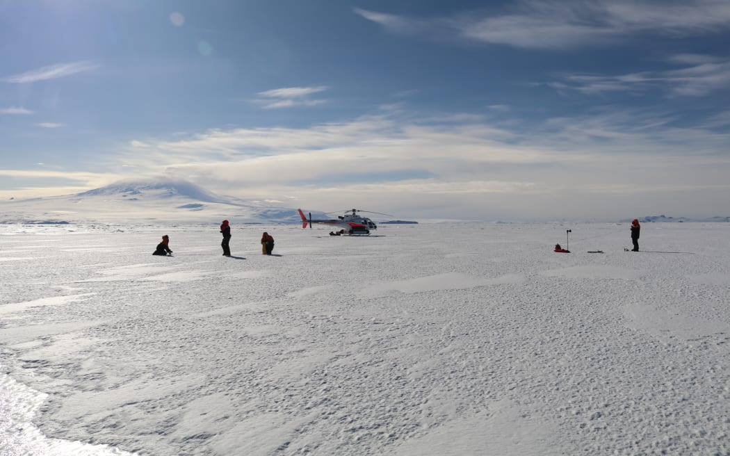 A wide shot of four figures either standing or kneeling on a white flat expanse of ice with a helicopter nearby. The sun is creating a lens flare in the blue sky, with a smattering clouds on the horizon, above a snow-covered hill.