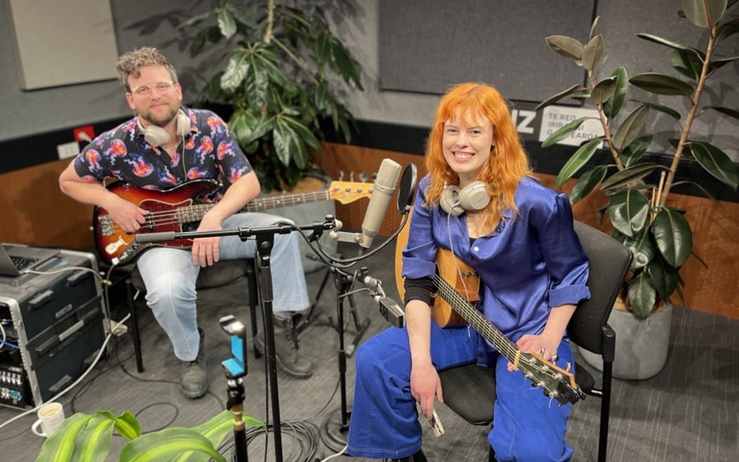 Emily is sitting with her guitar in front of a plant in the RNZ Christchurch studio. She has orange hear and is wearing a blue outfit. It is very eye catching, in a good way. To her left you can see her bass guitarist sat in front of another plant.