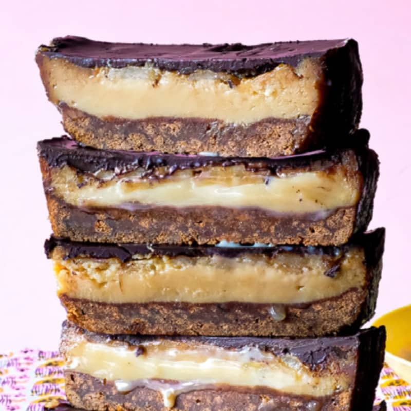 A vertical stack of five pieces of banana caramel slice, showing a cross-section of biscuit base, gooey caramel and chocolate topping, on a pink patterned cloth.