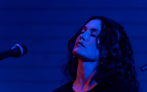 Babe Martin stands onstage under blue light.