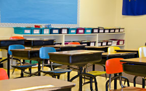 Students away from a classroom, which is filled with empty school desks.