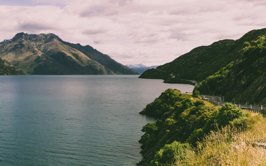 A road runs through scrub alongside a lake with mountains in the background and a cloudy sky.