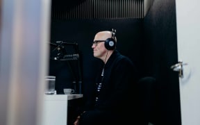 Garth Bray, TVNZ and Fair Go journalist, wearing headphones and smiling into a microphone in a small recording studio.