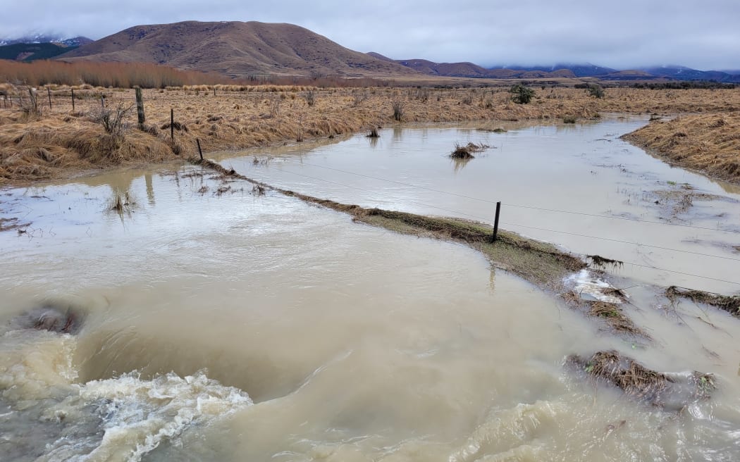 Brown floodwaters engulf a farm fence in a brown grassy flat, with mountains and low cloud in the background.