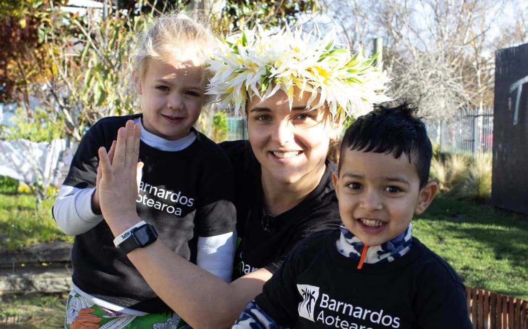 Amelia Kerr is wearing a Barnardos Aotearoa T-shirt and standing next to two smiling children.