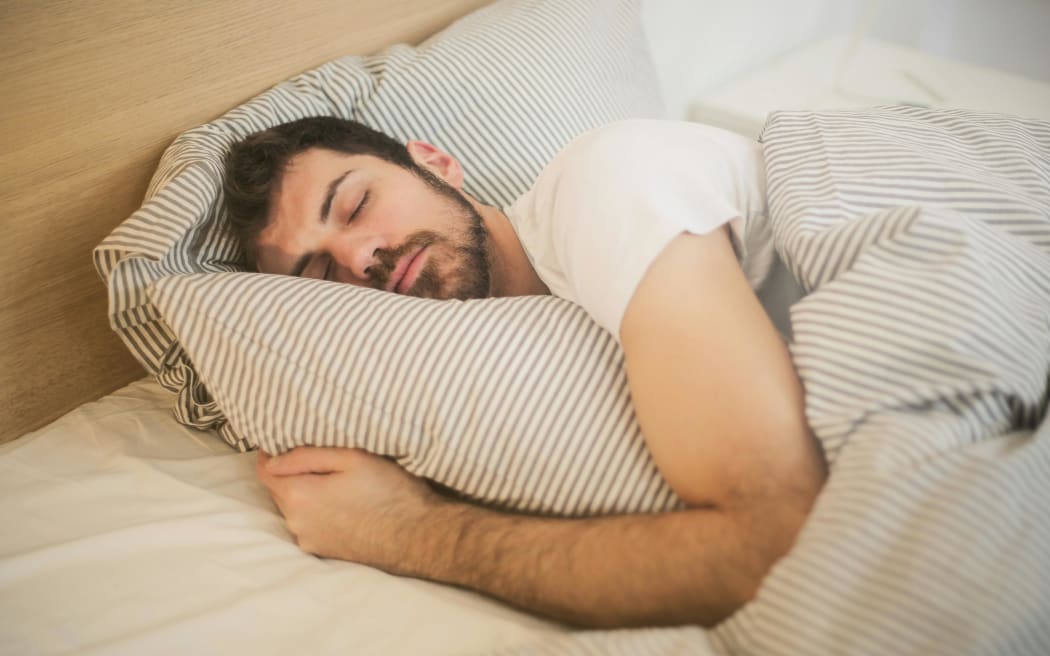 A bearded man with eyes closed hugs his pillow in bed.