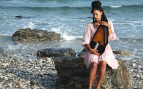 British folk musician Angeline Morrison sitting on a rock by the sea holding an autoharp.