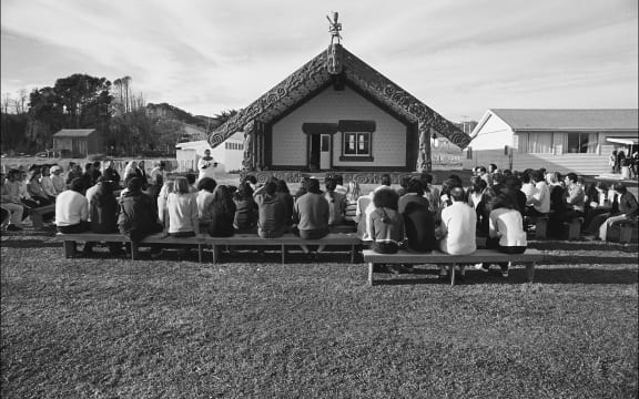 A black and white photo of a large group seated on benches outdoors, facing a marae.