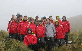 A group photo of some of the Sir Peter Blake Trust expedition team amongst the tussock on Col Lyall saddle. They are smiling, wearing red Blake jackets, with black Blake hats, or bright yellow hoods. Second to the right at the front is Jacob Anderson, who is wearing a grey jacket.