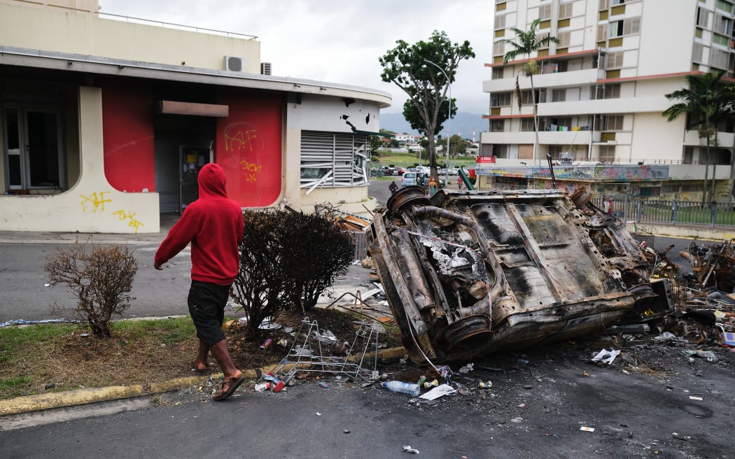 A man walks past a burnt vehicle at an independantist roadblock at Magenta Tour district in Noumea, France's Pacific territory of New Caledonia, on May 22, 2024. The Pacific territory of 270,000 people has been in turmoil since May 13, when violence erupted over French plans to impose new voting rules that would give tens of thousands of non-indigenous residents voting rights. The unrest has left six people dead, including two police, and hundreds injured. (Photo by Theo Rouby / AFP)