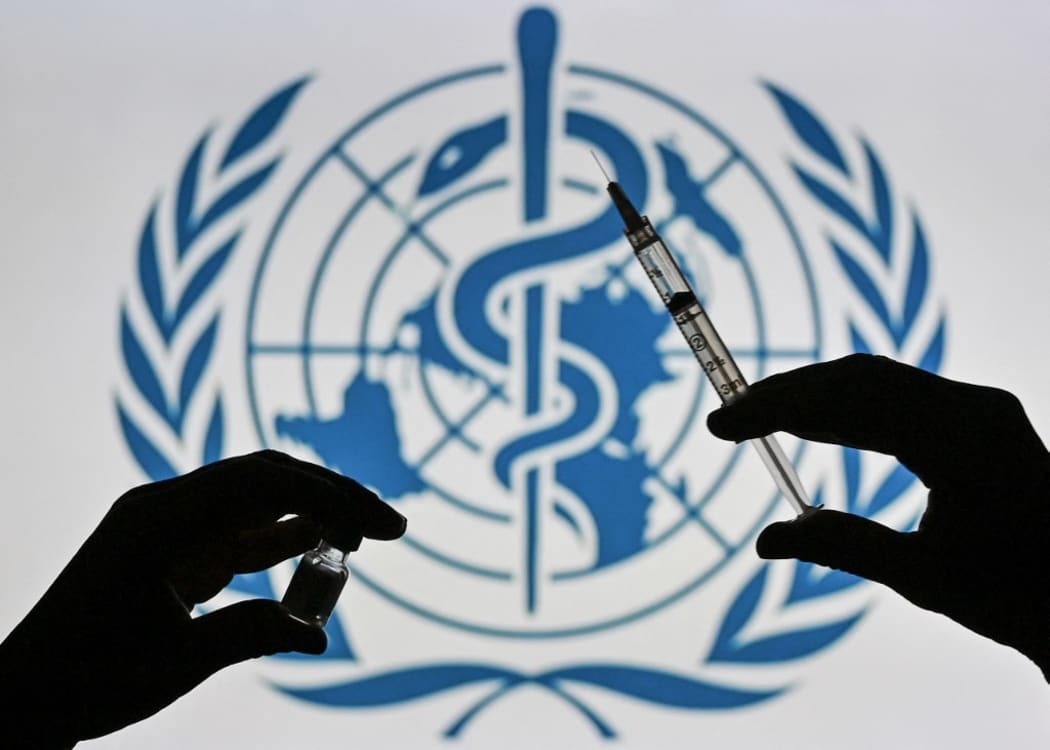 An illustrative image of a person holding a medical syringe and a Covid-19 vaccine vial in front of the World Health Organization logo displayed on a screen.