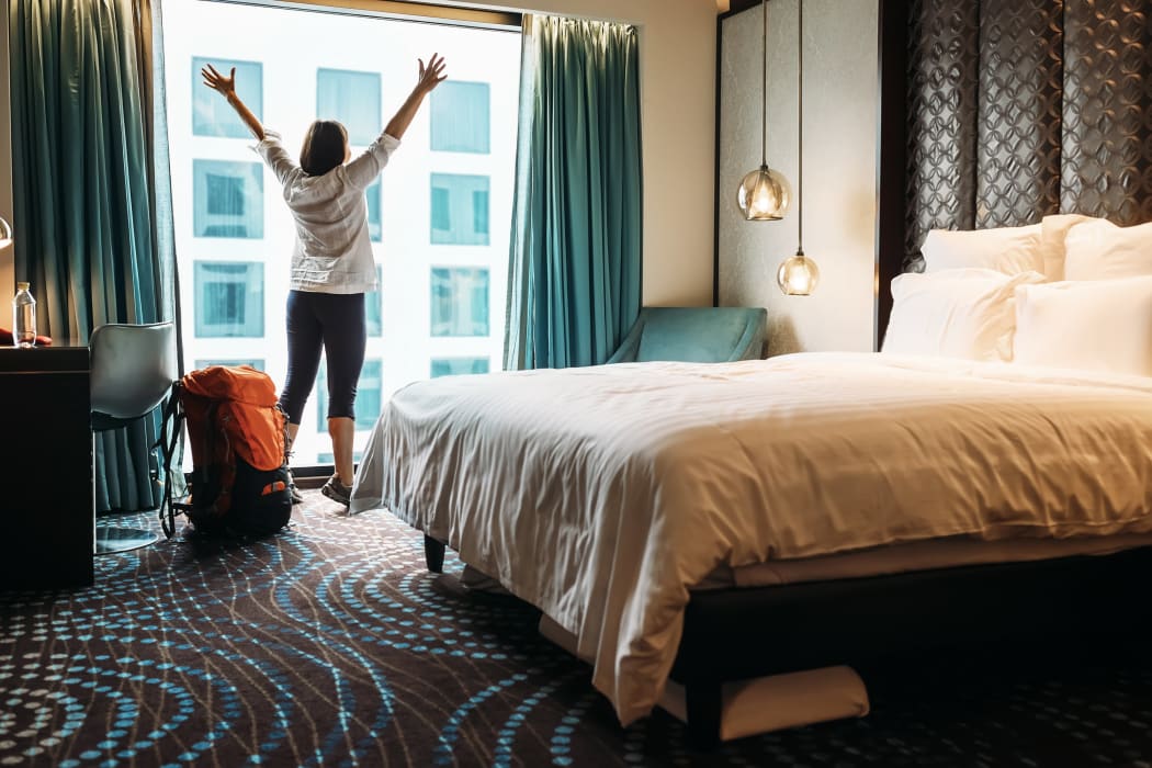 A woman backpacker stands next to her luggage and a window in a hotel room.