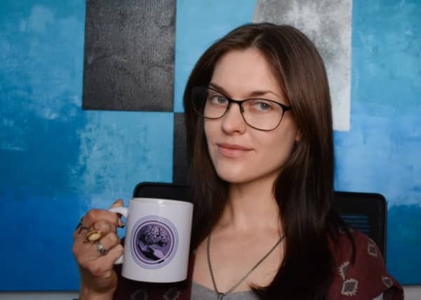 Rose Guingrich has long brain hair and thin framed optical classes and carries a mug.
