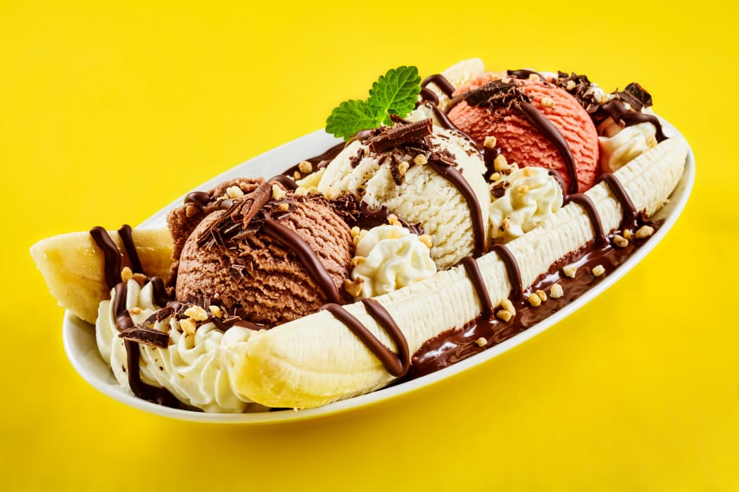 Tropical banana split with chocolate drizzle over three scoops of chocolate, strawberry and vanilla ice cream on fresh bananas, yellow background