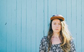 Person with long hair and cap standing in front of a blue painted wall