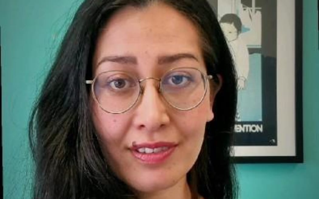 This is a headshot of Anita. She is in front of a green wall with a frame on it. It's not really possible to see what's in the frame because her head is in front of it. Anita has slightly over shoulder length black hair. She is wearing wire framed round glasses.