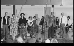Douglas Lilburn (fourth from righ, in checked jacket), Ashley Heenan (2nd from right on stage), and members of the Schola Musica at a special broadcast concert honouring Douglas Lilburn on his 65th birthday, in Symphony House, Willis Street, Wellington. November 1980.