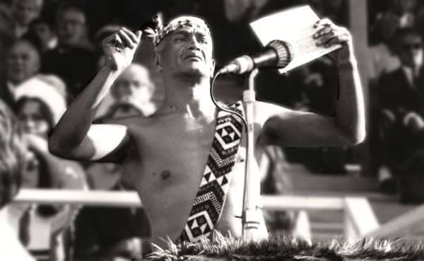 Bill Kerekere, wearing traditional Māori attire, holds a piece of paper and raises his arms.