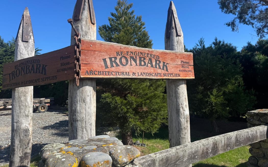 The entryway to Re-Engineered Ironbark, showing four large upright wooden poles capped with steel with a large wooden sign. In the foreground, a gateway made of riverstones