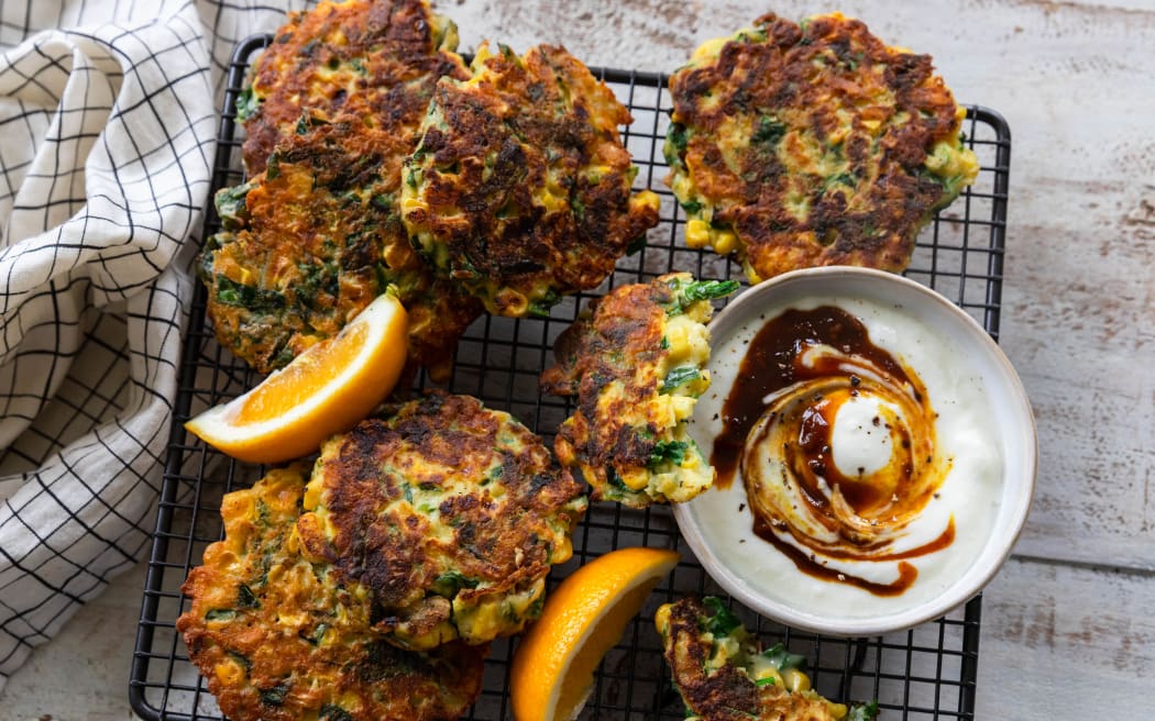 A wire tray topped with corn, green chilli, spinach and cheese fritters, garnished with lemon wedges and a bowl containing chutney swirled through yoghurt.