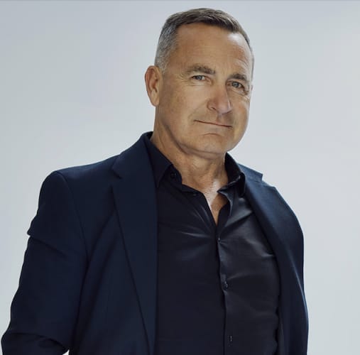 White man in shirt and blazer looking towards camera with a plain background