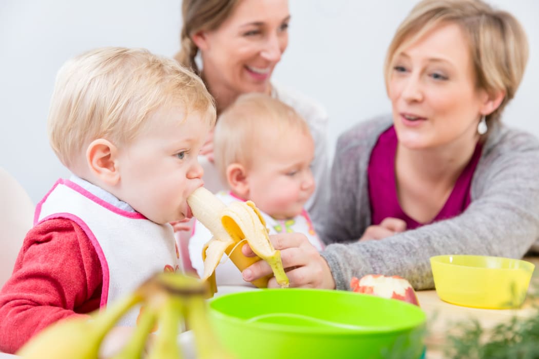 Portrait of a cute and healthy baby girl with blue eyes looking at her mother, while eating nutritious fruit puree next to another baby at home