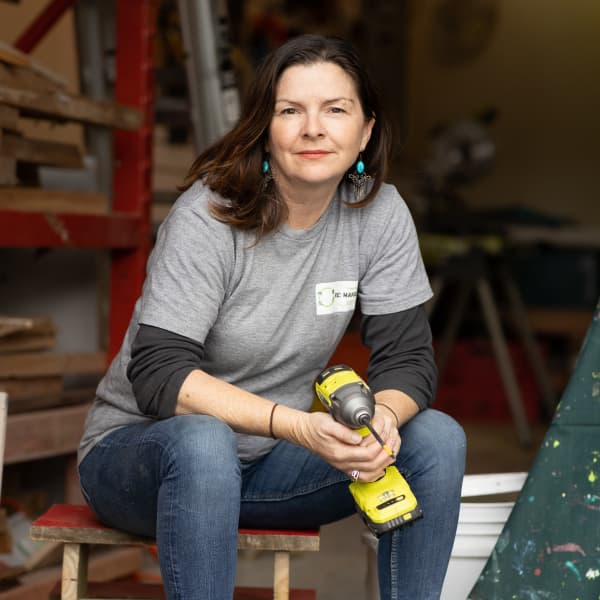 The ReCreators founder Ger Tew sitting on a stool in her woodworking garage, holding a drill.
