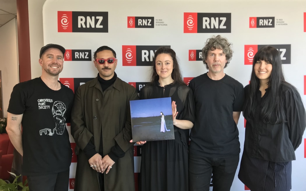 Five band members pose in front of a Radio New Zealand background. The woman in the middle wears a black dress and is holding up a