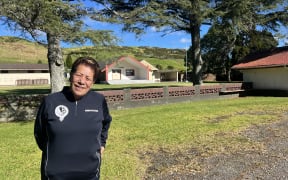 A portrait of Maybelle as she stands on some grass in front of a cream and red trimmed Marae flanked by trees, with a bright blue sky int he background.