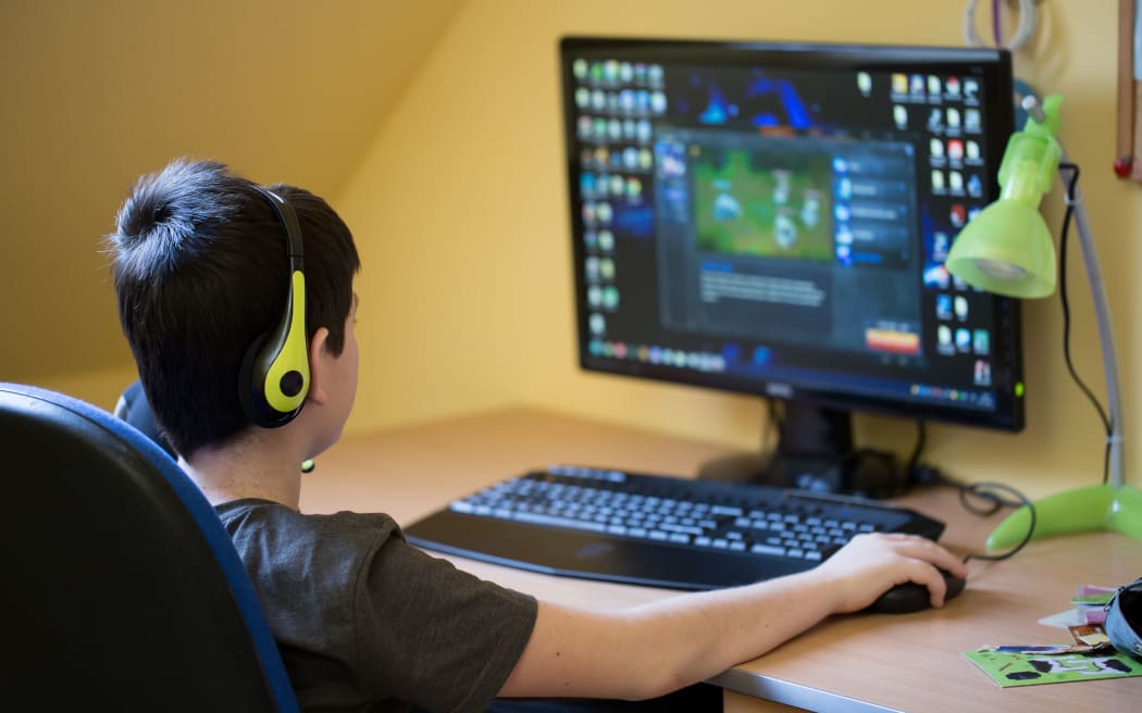 A photo of a teenager boy taken from behind. He is using computer at home with headphones to play a game in his room.