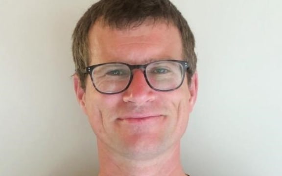 Diarmaid smiles at the camera. He is wearing glasses and a black medical uniform.