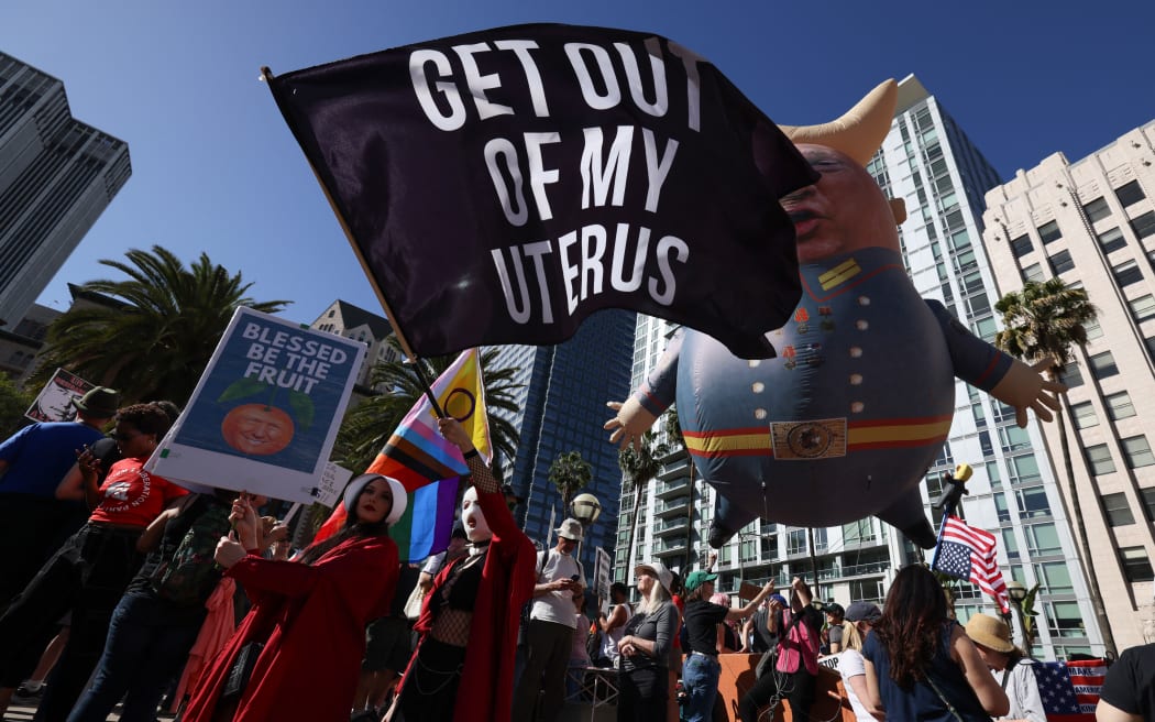 A large balloon with an image of US President Donald Trump floats above a protestor dressed as a character from 'The Handmaid's Tale' during the nationwide 'Hands Off!' protest against Trump and his advisor, Tesla CEO Elon Musk, in downtown Los Angeles on April 5, 2025. Protesters flooded the streets of several major US cities on Saturday to oppose the divisive policies of President Donald Trump, in the largest demonstrations since his return to the White House. Opponents of the Republican president's policies -- from government staffing cuts to trade tariffs and eroding civil liberties -- rallied in Washington, New York, Houston, Los Angeles and Florida, among other locations. (Photo by Etienne Laurent / AFP)