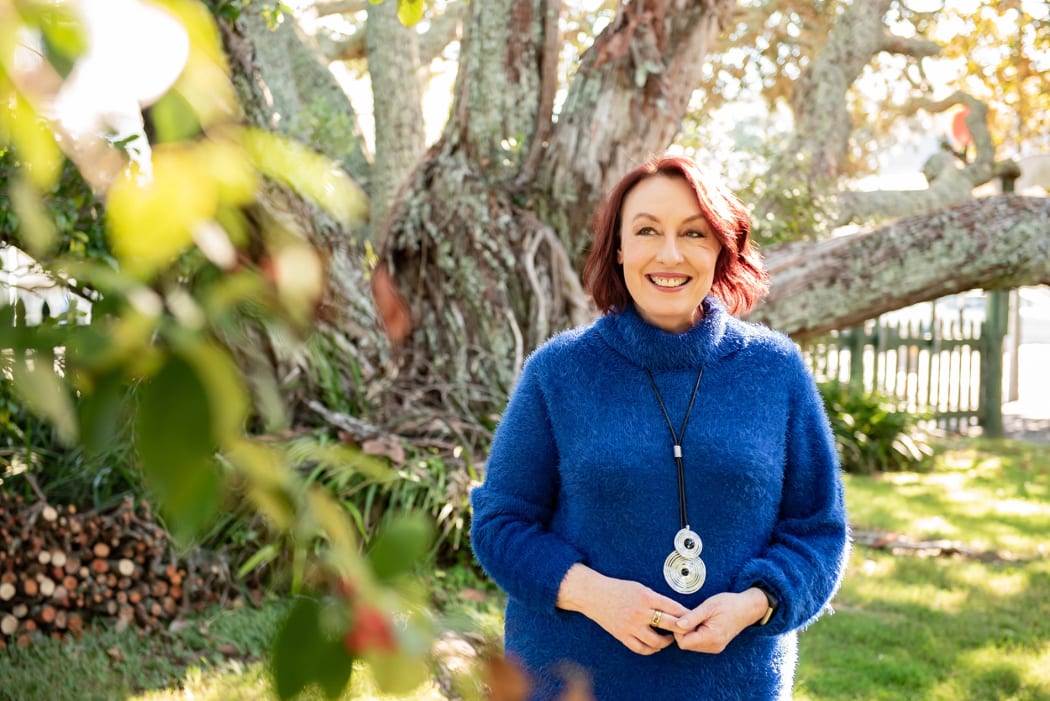 neuroscientist Christine Ozolins wearing a blue sweater stands in front of a large tree with some leave out of focus in the foreground