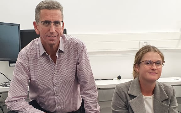 A man in a shirt and a woman in a blazer sit next to a large contraption with three holes in its surface.