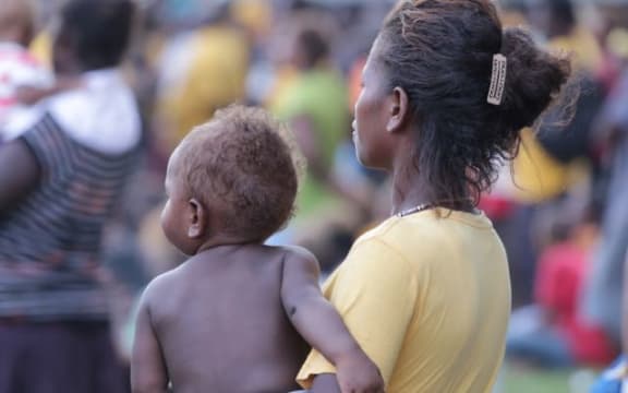 A woman voter nurses a child while listening intently to a campaign spiel at a political rally in Guadalcanal.
