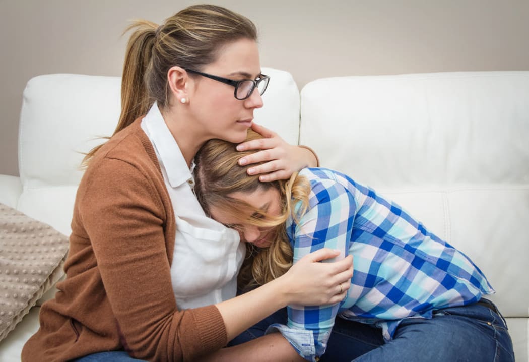 A photo of a mother on the couch embracing and comforting an anxious looking teenage daughter